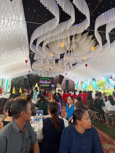 The scene is an outdoor event with many people seated at tables under a large tent-like structure, which is decorated with white fabric and hanging lights. There is signage that says, "Sự Kiện Tiệc Cuối Tuần Danh Tiến Danh" followed by a phone number. People are mostly seated and appear to be eating or talking. The location is likely Bình An, Vietnam.