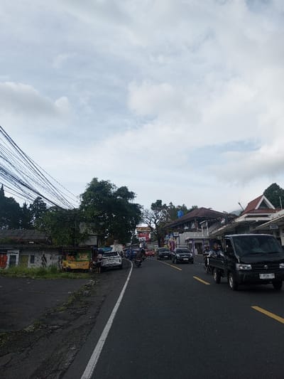 A street scene with light traffic on a cloudy day. A black Suzuki Carry pickup truck drives in the foreground. Other cars and motorcycles occupy the road. Buildings are visible on both sides, including one with a large "JOTUN" sign. Power lines dominate the left side of the frame. A food stall sits on the roadside.