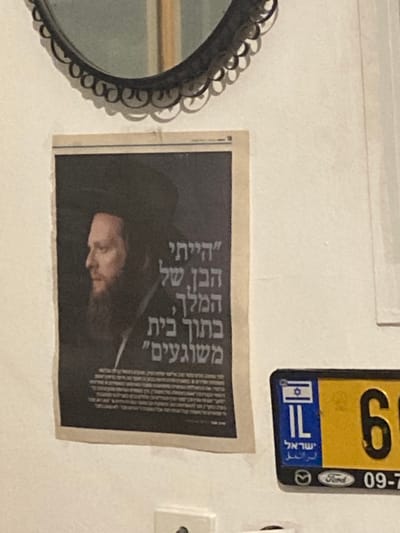 The image shows a white wall in a home in Kokhav Ya'ir Tzur Yigal, Israel. On the wall, there is a framed mirror in the upper left corner and a poster of a man with a black hat and beard below it. The man appears to be the subject of a newspaper article and the poster is likely an advertisement. There is also a yellow license plate attached to the wall in the bottom right corner. The license plate has a blue Israeli Star of David and reads “IL” in the upper left corner.