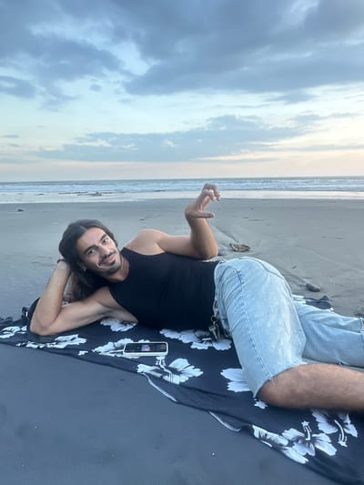 A man lies on a black and white patterned cloth on a sandy beach. He is facing the viewer, supported on his elbows and legs, with his right hand raised. He has long dark hair and a mustache. He wears a black tank top and light blue jeans. A cell phone rests on the cloth next to him. The beach stretches behind him, with the ocean and sky visible in the distance. The location is in the city of Kerambitan, Indonesia.