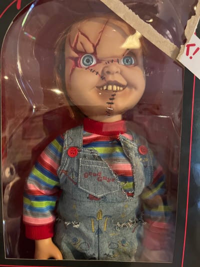 The image shows a close-up of a Chucky doll, a fictional character from the "Child's Play" horror film franchise, displayed within its packaging. The doll is wearing a red and multi-colored striped long-sleeved shirt and blue denim overalls with red buttons. The doll's face is rendered in a somewhat grotesque and damaged manner, with red stitches and scars visible on its head and mouth, and it has bright blue eyes. The doll is presented in a clear plastic box, which appears to be partially damaged or opened at the top, revealing cardboard material and a torn label with red markings. The background is dark with red accents, suggesting it is part of the product's packaging or display. The image does not contain any elements that would indicate a location in the city of Schwäbisch Hall, Germany.