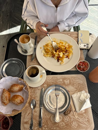 An overhead shot shows a partially eaten plate of potatoes and eggs, a small bowl of salsa, and a phone. A second place setting includes a fork, knife, and spoon atop a plate with napkins. A basket of bread sits beside cups of coffee and a drink. A person in a white jacket is visible at the edge of the frame. The meal is set on a table with patterned placemats and a black metal grate.