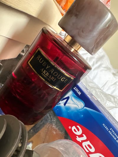 The image displays a cluttered, indoor scene featuring multiple consumer products and objects. Dominating the center is a ruby-red glass perfume bottle with a black label bearing gold text that reads "RUBY ROUGE AKBARI PERFUME." Its marbled grey cap is positioned slightly askew above the bottle's neck. To the right and behind the perfume bottle, a blue and white Colgate toothpaste box is partially visible, showing text such as "WHITENING" and "FOR WHITER TEETH CRYSTALS." In the lower left, a silver-toned metallic object with visible ridged components is present. Further background elements include stacked light beige material (possibly paper or fabric) in the upper left, an orange item in the upper right, and white, possibly plastic or fabric, beneath the toothpaste box. No individuals, specific actions, or interactions are depicted. The exact location in the city of Galway, Ireland, cannot be determined from the content of the image.
