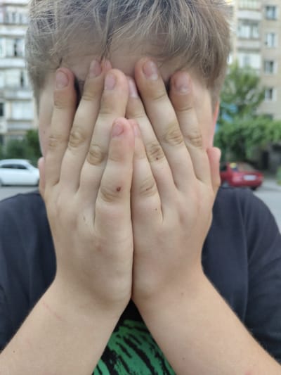 A medium shot captures a person from the chest up, with their face completely obscured by their hands. The individual's light brown hair is visible at the top of the frame. They are wearing a dark gray t-shirt, partially revealing a green and black patterned shirt underneath. The background suggests an outdoor urban setting, with a building that appears to be in the city of Melitopol, Ukraine. The building has multiple windows and a light-colored facade. A red car is parked in the distance, and a white car is visible to the left. The lighting is consistent with daylight, with shadows present. The overall impression is one of privacy or distress, as the person shields their face.