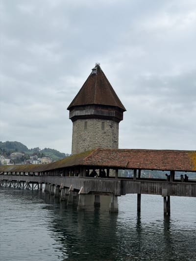 The image shows the Chapel Bridge (Kapellbrücke) and Water Tower (Wasserturm) in Lucerne, Switzerland, oriented vertically. The bridge, covered with a tiled roof, extends across the Reuss River. The octagonal Water Tower, constructed of stone, stands next to the bridge. A hillside with buildings is visible in the background. The sky is overcast, and the river's surface is rippled.
