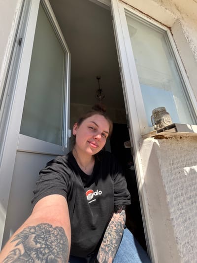 A person is leaning out of a window. The person is wearing a black t-shirt and has tattoos on their arms. The window is white and has a clear glass pane. The location is Chornomorskoe, Ukraine. The scene appears to be inside a building, looking out through the window. The wall around the window is white.