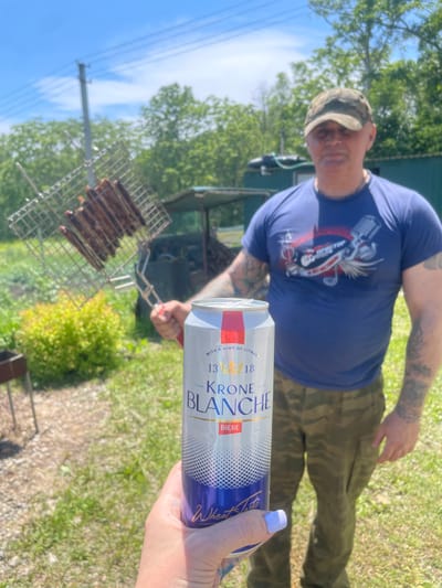 The image depicts an outdoor scene, likely in Yasnoe, Russia, with a person in the foreground holding a can of Krone Blanche beer. The can is white with blue and gold accents, and text indicating "BIERE" and "WITH A HINT OF CITRUS." Behind the beer can, a man wearing a camouflage baseball cap, a blue t-shirt with a graphic design, and camouflage pants stands in the middle ground. He is holding a metal grill basket filled with food, possibly sausages, over what appears to be a barbecue. The background shows a grassy area with trees and a green building, suggesting a rural or semi-rural setting. The lighting indicates it is daytime, likely sunny, with a clear blue sky.