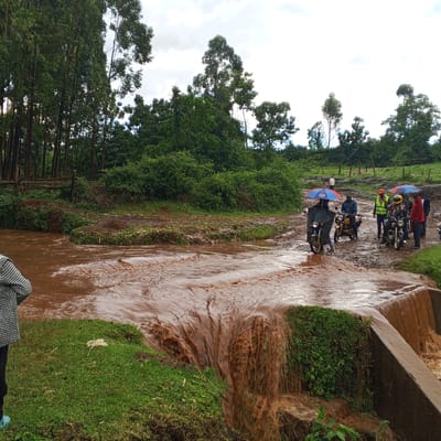 The image shows a flooded road in Keyian ward, Kenya. The water is brown and flowing rapidly over a concrete barrier. Several people are standing on the flooded road, including individuals on motorcycles. Some individuals are holding umbrellas, indicating rain or overcast weather. The surrounding environment features lush green vegetation, trees, and muddy terrain.