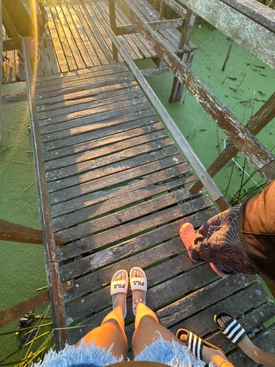 A wooden bridge spans a green, algae-covered body of water. Three people stand on the bridge. One wears dark pants and orange Crocs. Another has on black and white Adidas sandals. The third wears denim shorts and white Fila sandals. The bridge's wooden planks cast long shadows as sunlight streams across them. The structure shows signs of age with weathered wood and gaps between planks.