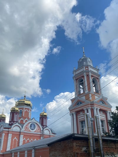 The image shows an exterior view of the Church of the Resurrection of Christ in Staraya Voskresenovka, Russia. The church features a prominent bell tower with a clock face and golden domes. The building is painted in shades of orange and white, with decorative white trim and columns. In the foreground, a brick wall and a metal roof partially obscure the lower portion of the church. The sky is blue with scattered white clouds. Overhead power lines are visible.