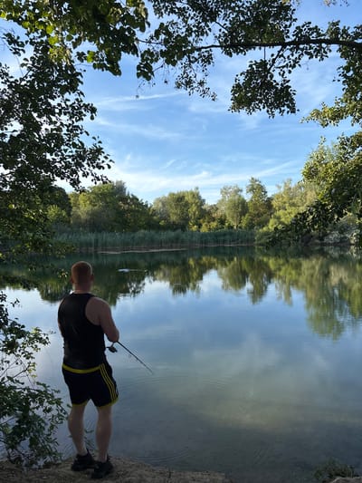 A man with red hair, wearing a black tank top and dark athletic shorts with yellow stripes, stands on the bank of a calm body of water, fishing. The man is viewed from behind, facing the lake. He holds a fishing rod, with the line extending into the water. Trees and reeds line the opposite bank of the lake, their reflections clearly visible on the water's surface. The sky above is blue with scattered white clouds. The foreground shows the man's feet and the rocky, earthy bank he stands on, with some greenery to the left. The scene appears to be a natural outdoor setting, likely a lake or pond. This location is identified as a pond in the municipality of Ludwigshafen am Rhein, Germany.