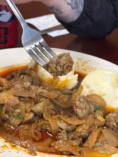 The image shows a plate of food consisting of mashed potatoes and a dish of cooked liver and onions. A fork holds a piece of the liver and onions mixture. The plate sits on a surface where a portion of what appears to be a red and black can is visible.
The image was taken in Des Plaines, United States of America.