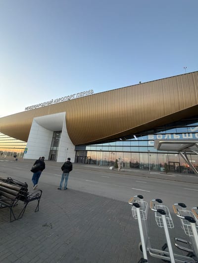 The image shows the front exterior of the Bolshoye Savino airport in Bolshoye Savino, Russia. The airport features a distinctive, curved facade with horizontal lines. The name of the airport is written in Cyrillic letters above the entrance. Two individuals are walking on the sidewalk in front of the building. A row of luggage carts is on the right side of the image. There is also a black metal bench. The sky is visible.