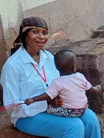 A woman with a patterned headscarf smiles and holds a baby in her lap. She wears a white collared shirt with a red lanyard. The baby wears a pink shirt and patterned pants. The woman sits outside near a brick structure. The image is rotated, making it appear sideways.