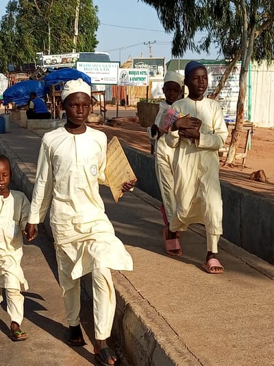 In Wuro Dole, Nigeria, three young boys in light yellow traditional attire walk along a paved walkway. The boy in the foreground, facing the camera, holds a wooden slate with Arabic script. To his right, another boy, wearing a blue cap, carries what appears to be a stack of colorful books or papers. The third boy, partially visible on the left, also wears the same yellow clothing and walks ahead of the boy with the slate. They are walking past a roadside market area with stalls covered in blue tarps. In the background, signs indicate businesses such as "MAHCOZY GUEST HOUSE LTD." and "MARQUIS HOTEL & APARTMENT." The environment is dusty and appears to be a sunny day with shadows cast on the walkway.