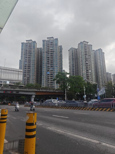 The image depicts a street scene in Nanhai District, China, under an overcast sky. In the background, multiple tall residential buildings with numerous windows dominate the skyline. In the middle ground, an elevated overpass with traffic lights is visible, with cars and motorcycles on the road below and on the overpass. The foreground features a multi-lane asphalt road with yellow and black striped bollards. Several vehicles, including motorcycles, cars, and a white van, are present on the road. Two individuals are seen riding motorcycles. Trees and greenery are interspersed among the buildings and along the overpass.