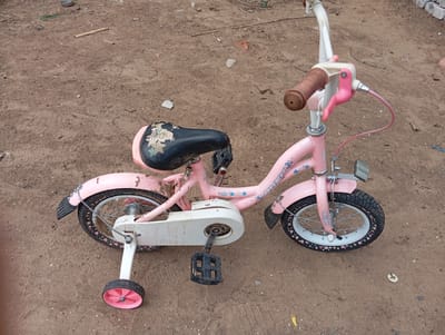 A pink children's bicycle is parked on a dirt surface. The bicycle has training wheels, a black seat with a cartoonish design, and pink handlebars with brown grips. The frame of the bicycle is adorned with light blue stars and hearts. The front wheel has a small reflector above it. The bicycle appears to be in a somewhat weathered condition, with some dirt and scuff marks visible on the frame and wheels. The background consists of dry, sandy soil with scattered small debris, suggesting an outdoor, possibly rural or undeveloped, location. The lighting indicates it is daytime. There is no visible text that is legible or relevant to understanding the scene beyond the branding on the bicycle. The overall impression is of a child's toy left outdoors.