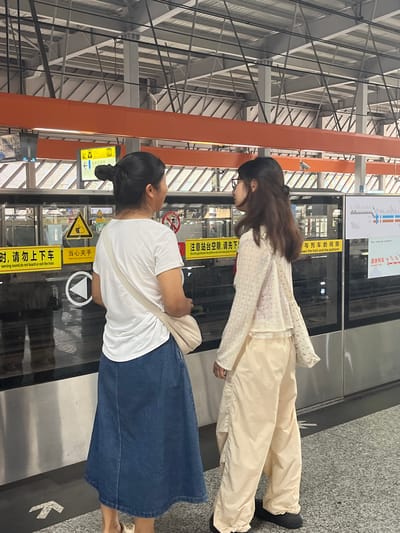 Two young women stand on a train platform in Tiaodeng, China, facing a train. The woman on the left wears a white t-shirt and a dark blue denim skirt, with her dark hair pulled up in a bun. She carries a cream-colored crossbody bag. The woman on the right has long, dark hair and wears a light-colored, long-sleeved open-knit cardigan over a cream-colored top and loose-fitting cream pants. She is wearing black shoes. They are standing near a train door, with safety signs visible on the glass doors. The platform is well-lit and appears to be a modern subway or train station, with the train's metallic exterior visible. The ceiling of the station is a complex metal structure. Yellow safety signs with Chinese text are affixed to the platform doors, warning passengers about train proximity and to mind the gap between the train and the platform.