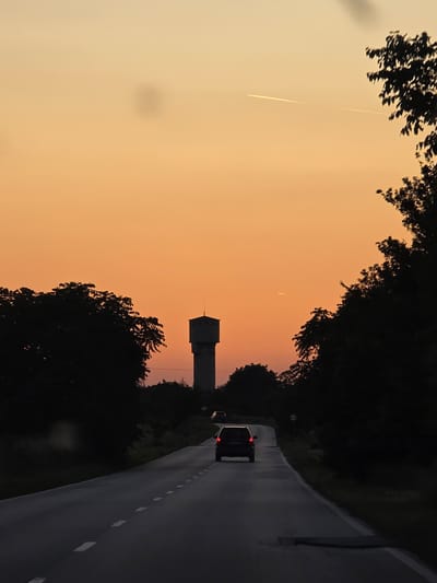 The image shows a road with two cars driving away at dusk. The sky is a gradient of orange, and trees line both sides of the road. A water tower is visible in the distance. The location is Karadjovo, Bulgaria. A vapor trail is also visible in the sky.