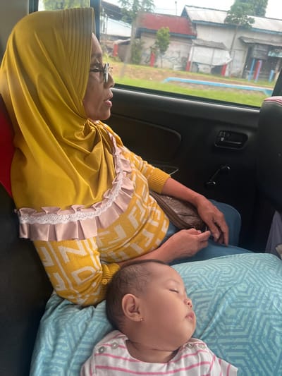 An elderly woman wearing a yellow headscarf and a patterned yellow sweater is seen in the backseat of a car. She is lying down with her eyes closed. A baby, wearing a pink and white striped shirt, is sleeping on a blue patterned pillow that rests on the woman's lap. The car window shows buildings and greenery outside. The interior of the car is visible, including a black door panel.