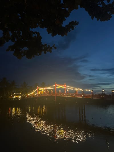 This media file depicts a night scene in Hoi An, Vietnam, showcasing a beautifully illuminated bridge spanning a body of water. The bridge, with its distinctive red architectural elements and an array of warm, yellow string lights, is the central focus. The lights create a shimmering reflection on the dark, rippling water below, adding a magical quality to the scene.

The time of day is clearly night, with a deep blue sky visible in the upper left portion of the image. Faint clouds streak across the sky, adding texture to the darkness. In the foreground on the left, the silhouetted branches and leaves of a large tree frame the view, adding depth and a sense of being outdoors. Palm trees are visible along the water's edge, subtly hinting at a tropical or subtropical location.

The overall mood conveyed is serene and enchanting, with the soft glow of the lights creating a romantic or festive atmosphere. There is no discernible activity of people or specific events taking place, suggesting a general ambiance of a peaceful evening. The location cues, such as the bridge design and the presence of palm trees, along with the explicit location context, point to Hoi An, Vietnam, a city known for its ancient town and charming waterways. No visible text is present in the image.