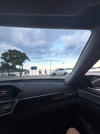 The image is a view from inside a car, looking out through the windshield. The sky is cloudy and appears to be late afternoon or early evening. In the distance, there is a body of water, likely the sea, with a coastline and hills beyond. A paved walkway runs along the water's edge, and several people are walking on it.
To the left, a large tree with a full canopy stands near the water. A blue road sign is visible on the walkway, indicating directions to "N. Ierissos," "N. Iraklitsa," "Kokkino," and "Kavala." To the right, a white Fiat 500 car is parked on the walkway. A street lamp is present near the car.
The interior of the car is visible in the foreground, showing the dashboard with a media display that reads "FM 107.0 MHz" and "KAVALA." The car's side mirror is also visible on the right. The overall scene suggests a coastal town in Greece, specifically Nea Peramos, given the road sign. The people are casually dressed and appear to be enjoying an evening stroll.