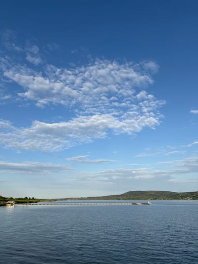 A wide-angle, horizontal shot captures a serene body of water under a partly cloudy, bright blue sky. The water occupies the lower half of the frame, displaying gentle ripples and a deep blue hue. In the middle ground, a long, white pier stretches across the water from left to right. On the far left, a yellow and white object, possibly a boat or a floating structure, is docked near the shore. The shore itself is lined with lush green vegetation. In the distance, a rolling, tree-covered hill dominates the horizon, with a smattering of buildings visible on its slopes. The sky above is a vibrant blue, adorned with scattered white clouds of varying sizes and densities, some appearing wispy and others more substantial. The overall impression is one of tranquility and natural beauty, likely a scenic spot in the vicinity of Казашко, Bulgaria, known for its water bodies and natural landscapes.