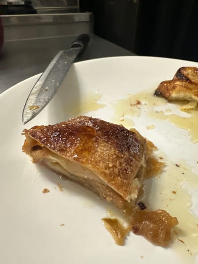 A close-up shot shows a partially eaten pastry dish on a white plate, accompanied by a knife. The pastry is golden brown with a flaky texture. Drippings of liquid surround the pastry remains. The knife has a metal blade and a black handle. The setting is a kitchen environment with stainless steel components visible in the background. There is no evidence of the image being taken in the city of Новоглаголево, Russia. There are no people or interactions visible in the image.