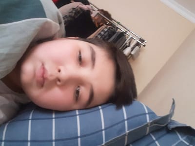 A young child lies on a blue plaid pillow, gazing languidly toward the viewer. Soft, rounded features and dark hair frame a serene face. To the left, a wall, and beyond, a clothing rack. The child's shirt, a light green and white striped garment, is partly visible. The shot is close-up, focusing primarily on the child's face.