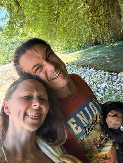This is a close-up, low-angle, outdoor selfie of a man and a woman. They are positioned close together, with the man's face on the left and the woman's on the right. Both are looking towards the camera with exaggerated expressions, scrunching up their faces and showing their teeth as if laughing or making a funny face.
The man has dark, curly hair and a beard, and is wearing a rust-colored t-shirt with a graphic design. The woman has long, dark brown hair and is wearing a patterned top.
The background shows a river on the left and a rocky shore with greenery on the right, suggesting they are by a body of water in a natural setting. The sunlight appears bright, indicating it is daytime.
On the man's t-shirt, there is visible text in a yellow, stylized font. The words "ENJOY" are prominent, and below that, "ADVENTURE" and "ON THE ROAD" are partially visible, along with "COUNTRY."
The overall mood is playful and intimate, capturing a moment of shared amusement between the couple in an outdoor environment. The location context suggests they are in Abgara, Abkhazia.