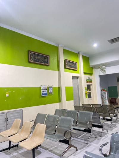 The image depicts a waiting area with rows of metallic chairs and some beige plastic seats. The room has a striking green and white striped wall, with two framed calligraphic artworks hanging on the green sections. One artwork is positioned higher on the wall than the other. Above the artworks, there is a ceiling with a ventilation grille and a light fixture. On the wall, above and between the two artworks, are several plastic-encased notices. A white wall fan is mounted on the upper left wall. To the right, a series of interconnected metallic chairs are arranged, with some beige plastic seats interspersed. The floor appears to be tiled. The scene suggests an indoor waiting space, possibly in a public building or clinic, given the arrangement of seating and the informational notices. There is no visible activity or people in the image. The lighting suggests it is daytime, or that the interior lights are on. The overall impression is one of an organized and clean waiting area.