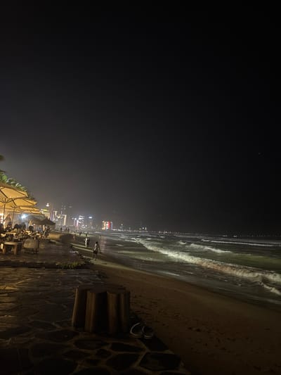 The image depicts a nighttime scene at a beachfront in Ngũ Hành Sơn Ward, Vietnam. The foreground features a dark-colored irregular stone-paved walkway or patio area. On this pavement, a cluster of three cylindrical wooden stools or bollards is visible, alongside a pair of dark-colored flip-flops. To the left, an outdoor dining or lounge area is established, characterized by multiple light-colored, open patio umbrellas illuminating tables and chairs where several indistinct figures are seated, appearing to socialize or dine. Behind this area, some dark vegetation, possibly palm trees, is discernible.
The mid-ground transitions to a sandy beach extending horizontally, where three to four individuals, likely children or young adults, are observed walking or standing near the waterline. To the right, the dark ocean with illuminated breaking waves rolls onto the shore. The background displays a long stretch of distant city lights along the coastline, indicating a densely built urban area with visible high-rise buildings. The sky above is dark and appears slightly hazy, with no clear celestial bodies visible. Artificial lighting from the establishments and distant city sources casts illumination across parts of the beach and waves.