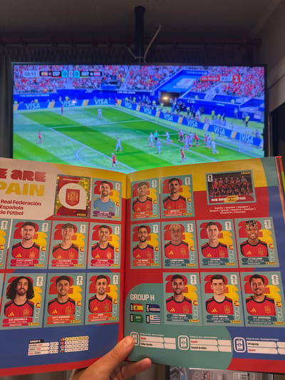The image shows a close-up of an open sticker album for the FIFA World Cup. The album is laid flat in front of a television screen displaying a soccer match. The album pages feature various soccer players' stickers, predominantly from the Spanish team, with their names, positions, birthdates, heights, weights, and club affiliations. The television screen shows a live soccer game with the score ESP 0-0 AUT displayed. The background behind the TV is out of focus, but appears to be a room interior. The album itself is a physical object, with glossy pages and printed stickers. The lighting in the scene is from the television screen and ambient room light. The camera is positioned above and to the right of the album, looking down at an angle. The image does not contain any information about the city of Prisovo, Bulgaria.