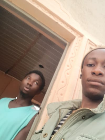 Two African men are posing for a selfie, possibly at the city of Dutsinma A, Nigeria. The man closer to the camera is wearing a green jacket with a black and white striped shirt underneath. The second man, positioned behind and below, is wearing a light blue tank top. They are standing in a doorway, the doorframe is light orange and has a decorative pattern. The interior behind them seems to be a metal-roofed structure.