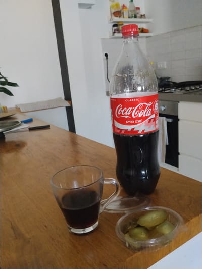 A half-full plastic bottle of Coca-Cola Classic stands upright on a wooden table. Next to it, a clear glass contains dark liquid, likely Coca-Cola. To the right of the glass, a small, clear plastic container holds several sliced pickles. The table surface shows wood grain. In the background, a kitchen counter with a stovetop and cabinets is visible. The wall behind the table is white, with a dark vertical stripe on the left. The overall scene appears to be an indoor residential setting, possibly a dining area adjacent to a kitchen. The image was taken in Be'er-Sheva, Israel.