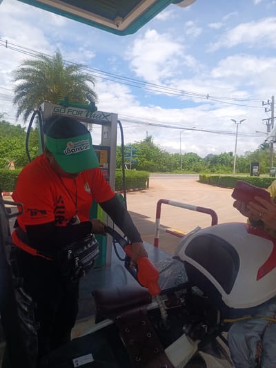 The image shows a gas station in Plai Phraya, Thailand. A gas station attendant wearing an orange shirt, black sleeves, a green visor, and a black fanny pack is using a red gas pump nozzle to fill the tank of a motorcycle. A person wearing a helmet and holding a red wallet or small purse is sitting on the motorcycle. The sky is blue with scattered clouds, and there are trees and a road in the background. The gas station has a sign that says "GO FOR Max" and a price display.