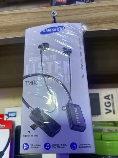 The image displays a retail product, specifically a Samsung wireless headset, packaged in clear plastic and presented on a shelf. The headset appears to be black with silver accents and is designed for activities such as exercise, listening to music, and making calls, as indicated by the text on the packaging. A portion of the packaging also highlights features like "Dual-device Connectivity," "Support TF card," and a battery life of "25-hr Long Playtime" with a quick charge option of "Charge 10-min, Play 100-min." The product is displayed against a wooden shelf, with other items visible in the background, including what appears to be computer hardware. The lighting suggests an indoor setting, likely a store, with no indication of time of day or weather. The overall impression is of merchandise ready for sale in a retail environment.