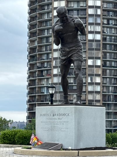 The image depicts a bronze statue of boxer James J. Braddock, known as the "Cinderella Man," in North Bergen, United States of America. The statue stands on a grey stone pedestal, which is inscribed with Braddock's name, nickname, and boxing achievements. In front of the pedestal, there is a small American flag and a bouquet of flowers placed on a dark plaque. To the left of the statue, a street lamp is visible. Behind the statue and pedestal, a tall, modern apartment building with numerous balconies and windows dominates the background. Trees and green foliage are present in the foreground and midground, providing a contrast to the urban architecture. The sky is overcast, suggesting a cloudy day. The scene appears to be a memorial site, likely in a park or public space, commemorating the boxer.