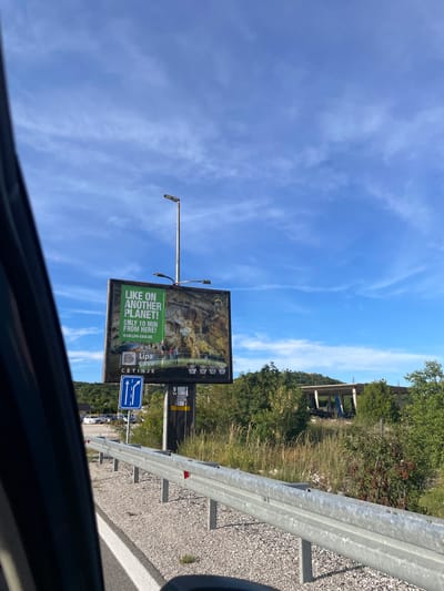 The image shows a roadside billboard in Očinići, Montenegro, advertising Lipa Cave. The billboard features an image of a cave interior with a group of people, and text that reads "LIKE ON ANOTHER PLANET! ONLY 10 MIN FROM HERE! WWW.LIPA-CAVE.ME" along with "Lipa Cave" and "CETINJE". Below the billboard, a blue road sign with white arrows indicates two lanes for going straight. A silver guardrail runs along the side of the road, which is covered in gravel. In the background, there are green trees and a partially constructed building. The sky is blue with wispy clouds.