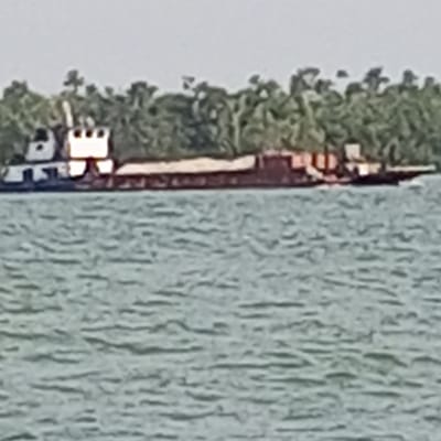The image depicts a large vessel, identified as a tugboat pushing a barge, navigating a waterway in Koko, Nigeria. The tugboat, positioned at the rear-left of the ensemble, is white and dark-colored with a distinct, multi-level cabin structure. A long, dark-colored barge, possibly laden with indistinct brown-orange cargo elements, extends forward to the right. The vessel is in motion, evidenced by a slight wake. The foreground is dominated by rippled, light green-blue water. In the background, a dense line of lush green trees forms a distant landmass or riverbank under a pale, hazy sky. No individuals are discernible.