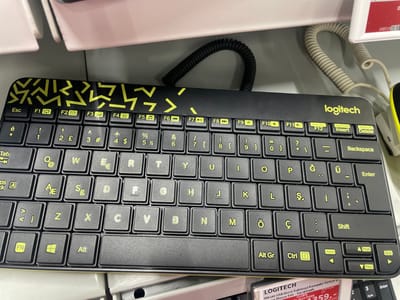 This media file shows a close-up, eye-level view of a black Logitech MK240 wireless keyboard with yellow geometric patterns. The keyboard is placed on a gray surface, likely a store display shelf, as indicated by the presence of price tags and other retail elements visible at the bottom of the frame. The setting appears to be a retail store in Bağcılar, Turkey, given the text "Bağcılar, Turkey" in the prompt.
The keyboard is the primary subject, with individual keys clearly visible. Prominent text includes the Logitech logo, function keys (F1-F12), alphanumeric keys, and special function keys like "Tab," "Shift," "Ctrl," "Alt," and "Fn." Several keys also feature Turkish characters such as "Ğ," "Ü," "Ş," "İ," and "Ç." The keyboard is connected by a coiled black cable.
Below the keyboard, partially visible, are parts of product packaging and price tags. One tag prominently displays "LOGITECH MK240 USB Alıcılı Kablosuz Kompakt Türkçe Mouse Seti - Sarı&Siyah" and "Uretim yeri: Çin" (Made in China). Another tag shows a price of "259,-" and a date "06.2026."
The lighting suggests an indoor, possibly well-lit retail environment, with no indication of time of day or weather conditions. The image focuses solely on the product and its immediate surroundings, with no people or animals present. The overall tone is neutral and informative, highlighting the product for sale.
