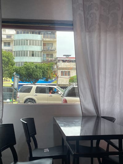 The image, taken from indoors through a window partly obscured by a patterned white curtain, shows an urban scene in Puerto La Cruz, Venezuela. Outside, there is a beige SUV with a person visible in the driver's seat, parked on a street lined with buildings of varying heights. One building has a light blue facade with white vertical stripes, and balconies with metal railings. Trees are interspersed along the street, partially obscuring the view of some buildings. A blue sign is visible on the street. Inside the room, a black table and chairs are partially visible in the foreground, suggesting the photograph was taken from a restaurant or indoor space. The lighting is subdued, with the outdoor scene appearing brighter than the interior.
