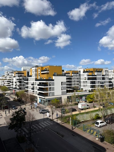 An aerial perspective depicts a modern urban scene in Rueil-Malmaison, France, under a clear blue sky with scattered white cumulus clouds. The primary subjects are contemporary residential and commercial buildings featuring white, grey, and yellow/ochre facades with numerous balconies.
In the midground, a street runs horizontally across the frame, with smaller intersecting streets. Several vehicles are present: a silver SUV parked on the right sidewalk, a dark-colored sedan partially visible on the far left, a silver sedan in the left lane approaching a crosswalk, and a white delivery van marked "Stricher Tripid" moving right-to-left in the center, near a "Grande Pharmacie de l'Arsenal" and "Laboratoire de biologie médicale" on the ground floor of a building.
Pedestrian sidewalks run alongside the streets. On the left side, three individuals are visible near a building entrance. Multiple bare deciduous trees and some evergreen trees line the streets and green spaces. A pedestrian crossing (zebra stripes) is prominent in the foreground. A green metal fence with an open gate section and yellow markings is visible on the right, alongside a white car and a dark waste bin. Streetlights, traffic signs (including a 30 km/h speed limit sign), and street markings are also present. Shadows indicate strong overhead sunlight.