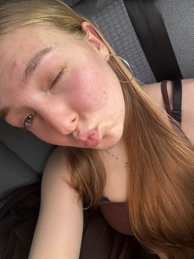 The image is a close-up, angled selfie of a young Caucasian woman with fair skin, light brown hair, and light-colored eyes. She is in what appears to be a vehicle, evidenced by a gray seat with a black seatbelt visible behind her. The woman is winking and puckering her lips, giving a kissy-face expression. She is wearing a thin silver necklace with a small pendant and large silver hoop earrings. Her right eye is partially visible, showing a light gray iris. She has some redness on her cheeks and a few small blemishes on her forehead. Her hair is long and light brown, falling over her shoulders. She is wearing a dark brown top. The background is a textured gray fabric, likely the car seat. There are no other individuals or animals visible in the image. The lighting suggests it is daytime, possibly taken with a phone camera. The image is in color. It is impossible to determine the specific location in the city of Kryukovo, Russia from the provided image.