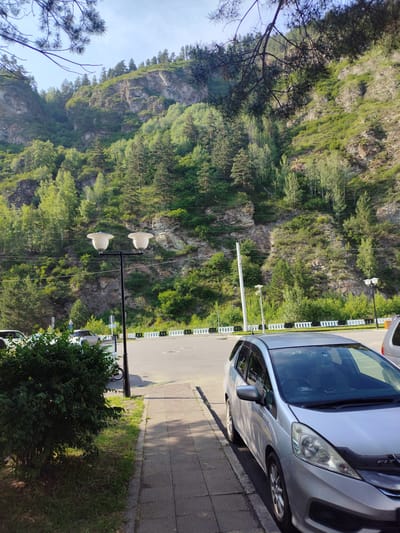 The image captures a serene daytime scene in Cheremushki, Russia, characterized by a dramatic mountainous backdrop. A sheer, rocky cliff face, covered in lush green trees and vegetation, dominates the upper half of the frame. The sunlight, suggesting it is either late morning or early afternoon, casts shadows and highlights the textures of the rock and foliage, creating a sense of depth.
In the foreground, a silver hatchback car is parked on a paved surface next to a sidewalk. The car appears to be a Honda Fit, with visible branding on the hood. Beside the sidewalk, a dense green bush adds a touch of natural color. A streetlamp with two white lamps stands between the bush and the road, and another similar lamp is visible further down the road. The road itself is paved and appears to be an asphalt surface, with a white barrier lining its edge. A few other cars are parked or driving on the road in the background, suggesting a public area or a street.
While the scene is mostly natural, the presence of the car, sidewalk, streetlights, and road indicates a developed area, likely a town or village situated near the natural landscape. No people are clearly visible in the main part of the image, though a faint figure might be present near the bushes in the distance. The overall mood is peaceful and tranquil, with the natural beauty of the mountains complemented by the human infrastructure. No discernible text is visible in the image.