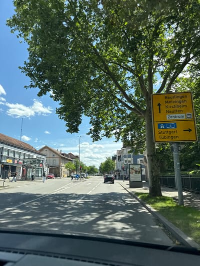 The image shows a street scene in Nürtingen, Germany. A yellow directional sign with German text indicates directions to Reutlingen, Metzingen, Kirchheim, Neuffen, Zentrum, Stuttgart, and Tübingen. A large tree with green leaves is prominently featured in the foreground, casting shadows on the road. On the left side of the street, there are buildings with shops and people walking. The sky is blue with scattered clouds. The road is paved and appears to be a multi-lane street with traffic markings. The overall scene suggests a sunny day in a moderately urban area.