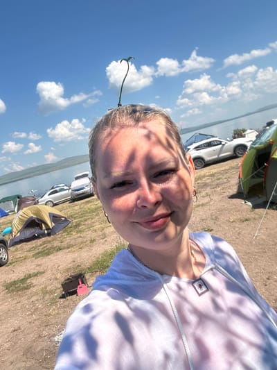 A young woman with blonde hair is taking a selfie outdoors. She has a small green sprout growing from the top of her head, which is bent over. Her face is turned towards the camera and she is smiling. She is wearing a light-colored hooded shirt with a tie-dye pattern. The background shows a body of water, a hilly landscape, and a campsite with several tents and cars. There are also shadows from trees dappling her face and shirt. The location is the city of Zhemchuzhny, Russia, specifically on a beach.