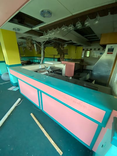 The image shows the interior of what appears to be a dilapidated building, possibly a former commercial space, within the Breckland District, United Kingdom. A pink and teal structure, potentially a counter or booth, dominates the foreground. Behind it, a partially visible kitchen area with stainless steel equipment is visible. The walls are painted yellow and light green. A rack holds rows of glassware, indicating the space may have been used as a bar or restaurant. Debris and construction materials such as wooden planks and white plastic sheeting, are scattered on the green floor. The building is in a state of disrepair. There are no people visible.