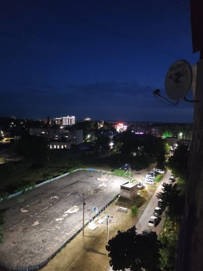 The image depicts a twilight view of a residential area in Kovrov, Russia. In the foreground, a dark, cracked asphalt surface, possibly a former sports field, is dimly lit by overhead lights. A blue fence borders the field, and beyond it, a grassy area and trees are visible. To the right, a paved road lined with trees curves through the scene, with several parked cars, including sedans and a white van. Further into the distance, apartment buildings are illuminated, with some appearing to be older, concrete structures and others more modern, with lit windows. A prominent building with a distinctive illuminated facade stands out among the city lights. The sky is a deep blue, with a few faint clouds visible. A satellite dish is attached to the side of a building in the upper right corner of the frame. The overall impression is of a quiet, nocturnal urban landscape.