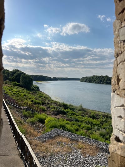 The image depicts a wide, calm river on a sunny day, viewed through an opening framed by weathered stone. The river stretches into the distance, bordered on the left by a dense line of green trees that form a dark, uniform edge against the water. On the right side of the riverbank, lush green vegetation grows abundantly, with patches of dry grass and a slope covered in grey gravel. A rusty metal staircase is visible descending through the green foliage, suggesting a path or access point.
The sky above is a clear blue, punctuated by scattered white clouds that appear fluffy and illuminated by the sun. The lighting suggests it is daytime, likely late morning or early afternoon, given the bright sky and the way the sunlight plays on the water and landscape. The overall scene conveys a sense of tranquility and natural beauty, with a rustic, somewhat unkempt feel due to the visible stone and gravel elements. There is no visible text in the image. The location context of Belene, Bulgaria, suggests this is a river landscape in that region, possibly near a dam or embankment structure given the man-made elements like the gravel slope and stairs.